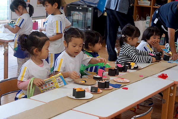 段ボールで作ったテーブルセンターのようなレールと、紙製の寿司でお寿司屋さんごっこをする子どもたち