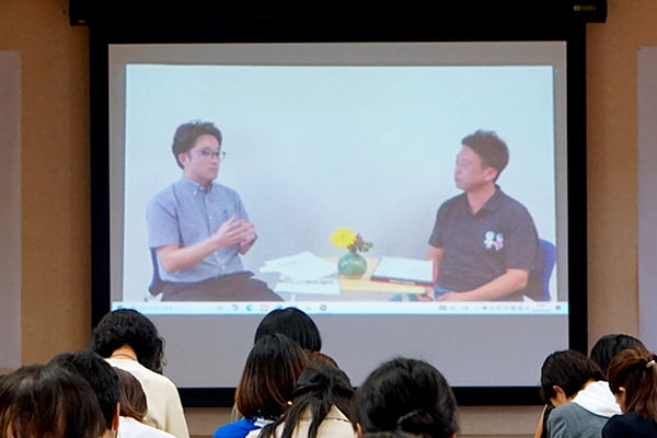 ステージ上のスクリーンに論文を読んでいる小・中学校の先生方の映像が映し出されている