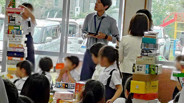 グループに分かれて様々な形や大きさの箱を重ねて身の回りのいろいろなかたちについて考える1年生算数科の公開授業とそれを見守る参観者