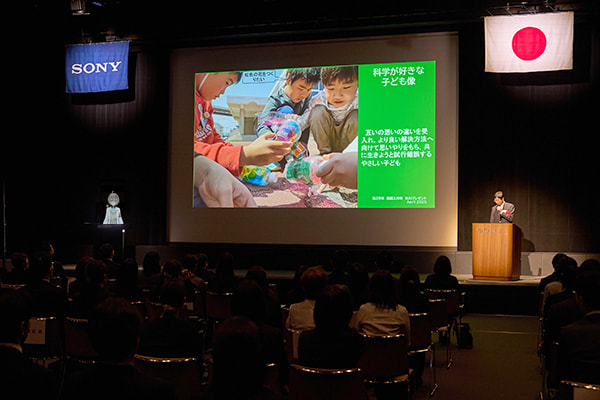 ステージのセンターに研究発表のスライドが映し出されている。上手演台に先生、下手演台に司会のかかぽさん。