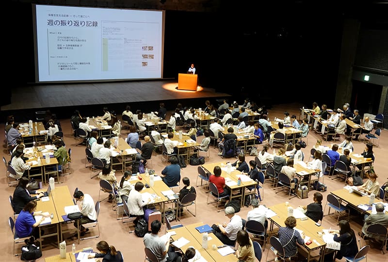 ホールでスクリーンに資料を投影しながら話す城崎こども園の先生と、その発表を聞いているたくさんの参加者